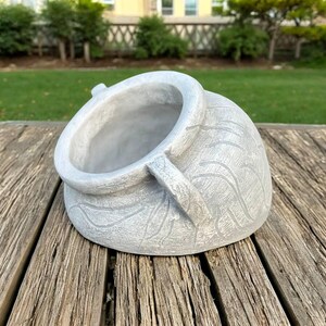 May include: A gray, cement-like planter with a rounded body and a wide opening. The planter has two small handles and a textured, carved design on the exterior. The planter is sitting on a wooden surface.
