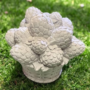 May include: A gray, decorative basket overflowing with artichokes. The artichokes are detailed with textured leaves and are arranged in a dense cluster. The basket has a woven design, and the sculpture is set against a backdrop of green grass.