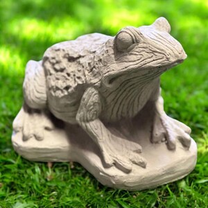 May include: A detailed, light gray frog statue with textured skin and realistic features. The frog is sitting on a small, flat base, with its legs and feet clearly defined. The statue is designed for outdoor garden decor.