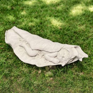 May include: A light gray stone sculpture of a sleeping baby wrapped in a blanket. The baby is lying on a leaf-shaped base, with detailed facial features and delicate hands and feet. The sculpture is placed on a bed of green grass.