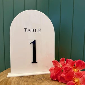 May include: White table number sign with the word "TABLE" and the number "1" in black. The sign is arched at the top and sits on a white base. Red flowers with yellow centres are to the right.