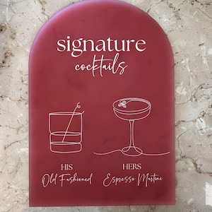 May include: A burgundy-coloured, arched sign with the words "signature cocktails" in white script. Below, line drawings of a cocktail and martini are labelled "HIS Old Fashioned" and "HERS Espresso Martini."