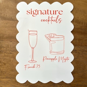 May include: White scalloped-edge sign with red text and illustrations. The words "signature cocktails" are at the top. Below are line drawings of a champagne flute and a cocktail glass, with the names "French 75" and "Pineapple Mojito".
