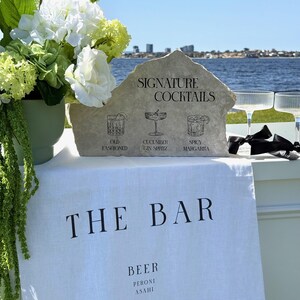 以下が含まれることがあります： 「THE BAR」の文字が入った白いリネンの布を使ったバーのセットアップ。石の看板には「SIGNATURE COCKTAILS」と飲み物のイラストが表示されています。左側には白と緑の花が入った花瓶があります。右側にはシャンパングラスが2つあります。