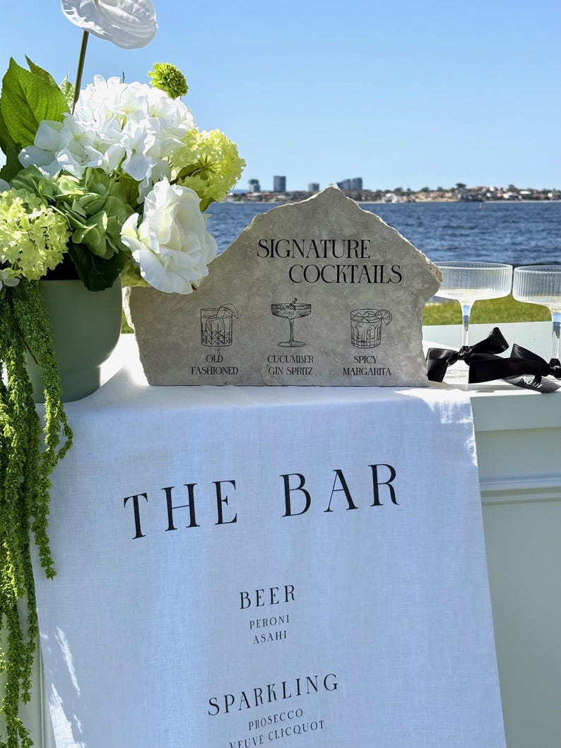 以下が含まれることがあります： 「THE BAR」の文字が入った白いリネンの布を使ったバーのセットアップ。石の看板には「SIGNATURE COCKTAILS」と飲み物のイラストが表示されています。左側には白と緑の花が入った花瓶があります。右側にはシャンパングラスが2つあります。