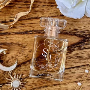 May include: A clear perfume bottle with a gold cap and a floral design, with the initials "S + M" in a white circle. The bottle is on a wooden surface, surrounded by gold jewellery and a white flower.