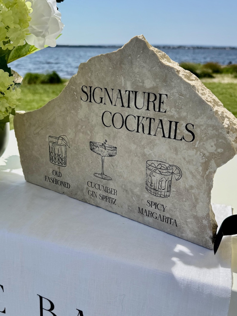 以下が含まれることがあります： 「Signature Cocktails」の文字と、Old Fashioned、Cucumber Gin Spritz、Spicy Margaritaの3種類のカクテルのイラストが描かれた石の看板。白いテーブルクロスの上に置かれ、背景には水と緑のぼやけた景色が広がっています。