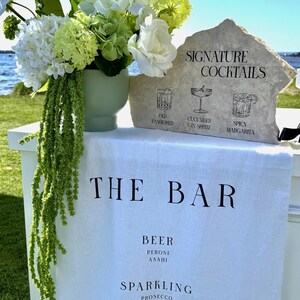 以下が含まれることがあります： 「THE BAR」と書かれた白いリネンの布があるバーのセットアップ。石の看板にはイラスト付きで「SIGNATURE COCKTAILS」と書かれています。白と緑の花が入った花瓶が近くにあります。バーではビール、スパークリングワイン、ワインを提供しています。
