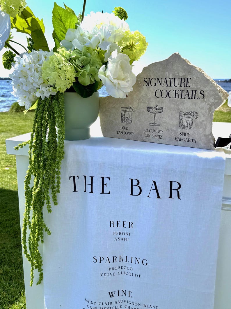 以下が含まれることがあります： 「THE BAR」と書かれた白いリネンの布があるバーのセットアップ。石の看板にはイラスト付きで「SIGNATURE COCKTAILS」と書かれています。白と緑の花が入った花瓶が近くにあります。バーではビール、スパークリングワイン、ワインを提供しています。
