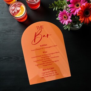 May include: An orange bar menu with the text "THE Bar" in red script. The menu lists bubbles, wine, and beer & cider options. Three cocktails with orange slices and a vase of pink and orange flowers are also visible.