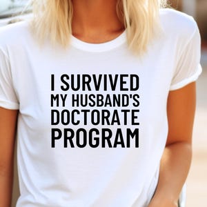 May include: White t-shirt with the text "I SURVIVED MY HUSBAND'S DOCTORATE PROGRAM" in a bold, black font. The shirt has short sleeves and a crew neck.