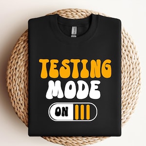 May include: A black crewneck sweatshirt featuring the text "TESTING" in yellow and "MODE" in white, followed by a white rectangular graphic containing the word "ON" in black and three vertical yellow bars, all in a rounded, bold font style.