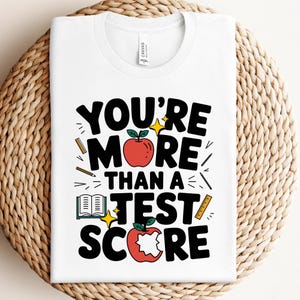 May include: A white t-shirt featuring the black text "YOU'RE MORE THAN A TEST SCORE" with graphic illustrations of two red apples, one with a bite mark, an open book, two rulers, a pencil, and yellow star accents printed on the front.