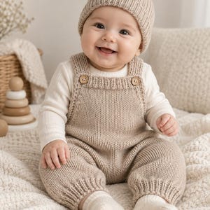 May include: A beige knitted overall with two wooden buttons at the straps, worn over a long-sleeved cream ribbed shirt. The outfit includes a matching beige knitted beanie with a pom-pom on top and cream-colored knitted socks on the feet.