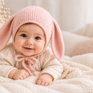 Peut inclure: Un bonnet tricoté rose pâle doux avec de longues oreilles de lapin tombantes et un lien à nouer sous le menton. Le bonnet est porté par un nourrisson souriant vêtu d'un haut à manches longues crème, allongé sur une couverture tricotée blanc cassé.