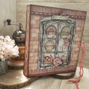 Puede incluir: Una caja decorativa con un diseño de ventana que presenta tazas y platillos. La caja tiene un patrón de ladrillos y una pancarta que dice "HOME SWEET HOME". Rosas rosas y una cinta rosa se suman a la estética vintage.