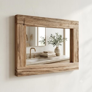 May include: A rectangular wall mirror featuring a thick, weathered natural wood frame with visible grain and texture. The mirror reflects a bathroom vanity with a white sink, a gold faucet, a stack of folded beige towels, and a vase with greenery.