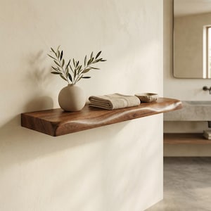 May include: A live edge wooden floating shelf mounted on a wall. On the shelf sits a round beige ceramic vase containing a green olive branch, a folded beige fabric towel, and a small, shallow, textured stone bowl in a neutral beige tone.