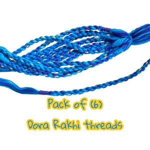 May include: A pack of six Dora Rakhi threads in shades of blue, with gold accents. The threads are braided and appear to be made of a soft, flexible material. The text "Pack of (6) Dora Rakhi Threads" is visible.