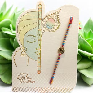 May include: A Raksha Bandhan greeting card with a colourful bracelet. The card features a gold illustration of a flute, peacock feather, and a face. The bracelet is made of twisted threads in red, blue, and yellow, with a gold charm.