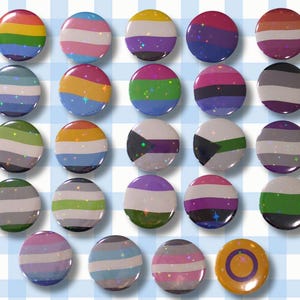 Könnte beinhalten: Eine Sammlung runder, farbenfroher Ansteckbuttons mit verschiedenen Pride-Flaggen-Designs. Die Buttons zeigen eine Reihe von Farben, darunter Regenbogen, Rosa, Blau, Gelb, Lila und mehr. Der Hintergrund ist ein hellblaues und weißes Karomuster.