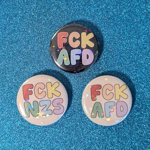 Könnte beinhalten: Drei runde Knöpfe mit rosa Hintergrund und glitzernder Oberfläche. Die Knöpfe haben den Text "FCK NZS", "FCK AFD" und "FCK AFD" in Regenbogenfarben.