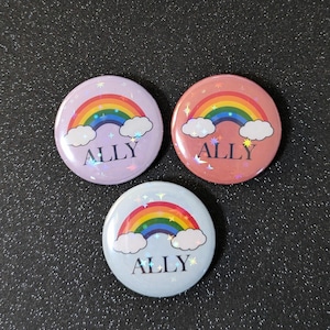 Könnte beinhalten: Drei Regenbogen-Button-Pins mit dem Wort "ALLY" in schwarzem Text. Die Buttons befinden sich auf einem dunkelgrauen Hintergrund. Die Buttons sind rosa, rot und blau.