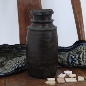 May include: A dark brown, cylindrical vase with horizontal grooves, on a wooden chair. A patterned textile with blue, black, and white designs is draped over the chair. Small, light-coloured, rectangular and circular objects are in front of the vase.