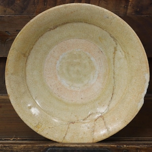 May include: A round, antique ceramic plate in light green and peach tones. The plate features concentric circles and visible cracks, indicating age and use. It rests on a dark wooden surface.