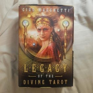 May include: A tarot card box featuring the artwork of Giro Marchetti. The box has a portrait of a woman with ornate headwear and jewelry. The title "Legacy of the Divine Tarot" is displayed in gold lettering. The background is a mix of warm colors.
