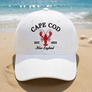 May include: White baseball cap with "CAPE COD" embroidered in black, a red lobster graphic, and "EST. 1602 New England." The hat is on a sandy beach with the ocean in the background.