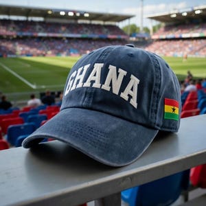 May include: A navy blue baseball cap with the word "GHANA" in white block letters. The cap features the Ghana flag on the side and is sitting on a metal railing in front of a stadium.