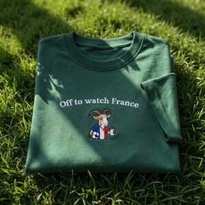 Puede incluir: Una camiseta verde oscuro con la frase bordada "Off to watch France". Debajo del texto hay una imagen bordada de una cabra con la bandera francesa, un bal&oacute;n de f&uacute;tbol y una taza. La camiseta est&aacute; doblada sobre hierba verde.