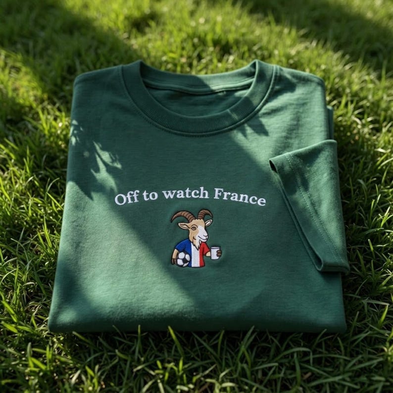 Puede incluir: Una camiseta verde oscuro con la frase bordada "Off to watch France". Debajo del texto hay una imagen bordada de una cabra con la bandera francesa, un bal&oacute;n de f&uacute;tbol y una taza. La camiseta est&aacute; doblada sobre hierba verde.