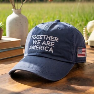 May include: Navy blue baseball cap with the text "TOGETHER WE ARE AMERICA" in white. The cap features a small American flag patch on the side. The hat is made of a soft material and has a curved brim.
