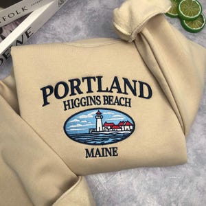 May include: Beige sweatshirt with embroidered text "PORTLAND HIGGINS BEACH MAINE". The design includes a lighthouse illustration in an oval frame. The sweatshirt is made of a soft material.