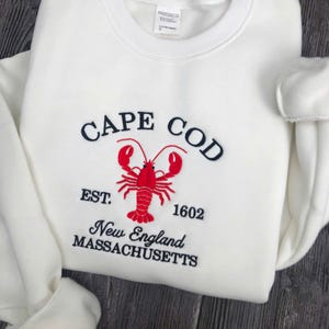 May include: White sweatshirt with the words "CAPE COD" arched across the top, a red lobster graphic in the center, and the text "EST. 1602 New England MASSACHUSETTS" below. The sweatshirt is a crew neck style.