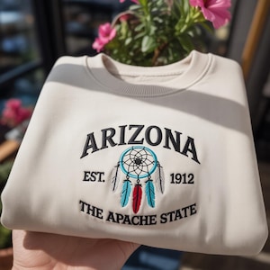 May include: Cream-colored sweatshirt with the word "ARIZONA" embroidered in black above a dreamcatcher design. The design includes the text "EST. 1912" and "THE APACHE STATE". The sweatshirt is folded.