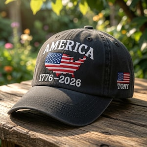 May include: A charcoal grey baseball cap featuring white embroidered text that reads AMERICA and 1776-2026. The front displays a red, white, and blue map of the United States. A small American flag and the name TONY are embroidered on the side.