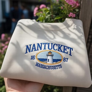May include: Cream-colored sweatshirt with the embroidered text "NANTUCKET" in blue. Below the text is an oval design with a lighthouse, the numbers "16" and "87", and the word "MASSACHUSETTS". The sweatshirt is folded.