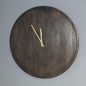 May include: A round, dark brown wooden clock with gold-colored hands. The clock has a simple, minimalist design and is mounted on a gray wall. The wood grain is visible, adding texture to the clock face.
