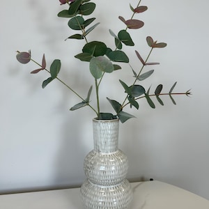 Peut inclure: Un vase en céramique blanc avec un design texturé empilé contient des branches d'eucalyptus. Les feuilles sont un mélange de vert et de rouge-violet, créant un arrangement naturel et décoratif. Le vase est posé sur une surface blanche effet marbre.