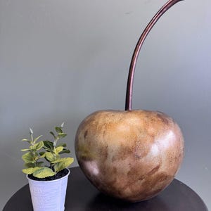 May include: A large, bronze-coloured cherry sculpture with a long, curved stem. The sculpture has a textured surface and sits on a dark, round table. A small white pot with greenery is to the left of the cherry.