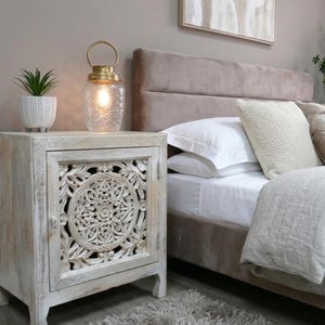 May include: A white-washed wooden bedside cabinet with a carved floral door design. A glass lantern with a gold handle and a small potted plant sit on top. The cabinet is next to a bed with white bedding and decorative pillows.