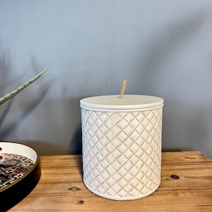 May include: A white, cylindrical container with a diamond-patterned exterior and a matching lid. A small, natural-coloured rope is attached to the top of the lid. The container sits on a wooden surface.