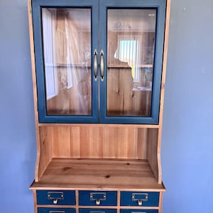 May include: A wall-mounted cabinet with a natural wood finish and navy blue accents. The cabinet features glass-paned doors, brass handles, and six small drawers with brass label holders. The cabinet is designed for storage and display.