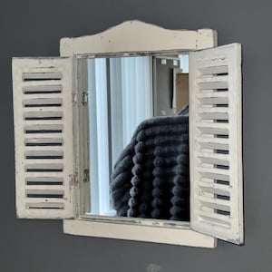 May include: A decorative wall mirror with a distressed white frame and shutter-style doors. The mirror reflects a room with a gray blanket. The shutters are open, revealing the mirror's surface.