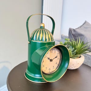 Può includere: Orologio a lanterna in metallo verde con manico e dettagli dorati. Il quadrante è bianco con numeri e lancette nere. L'orologio è su un tavolo marrone scuro.