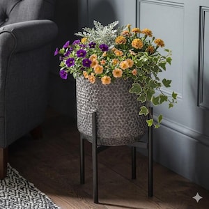 Op de afbeelding: Een gehamerde metalen plantenbak gevuld met levendige bloemen in paars, oranje en geel. De plantenbak staat op een zwarte metalen standaard. Hangende groene klimop voegt een vleugje frisheid toe. Een decoratief item voor de woondecoratie.