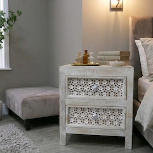 May include: A white, distressed wooden bedside table with two drawers, each with a floral cut-out design. The table has silver knobs and is next to a bed with neutral bedding. Decorative items are on top.